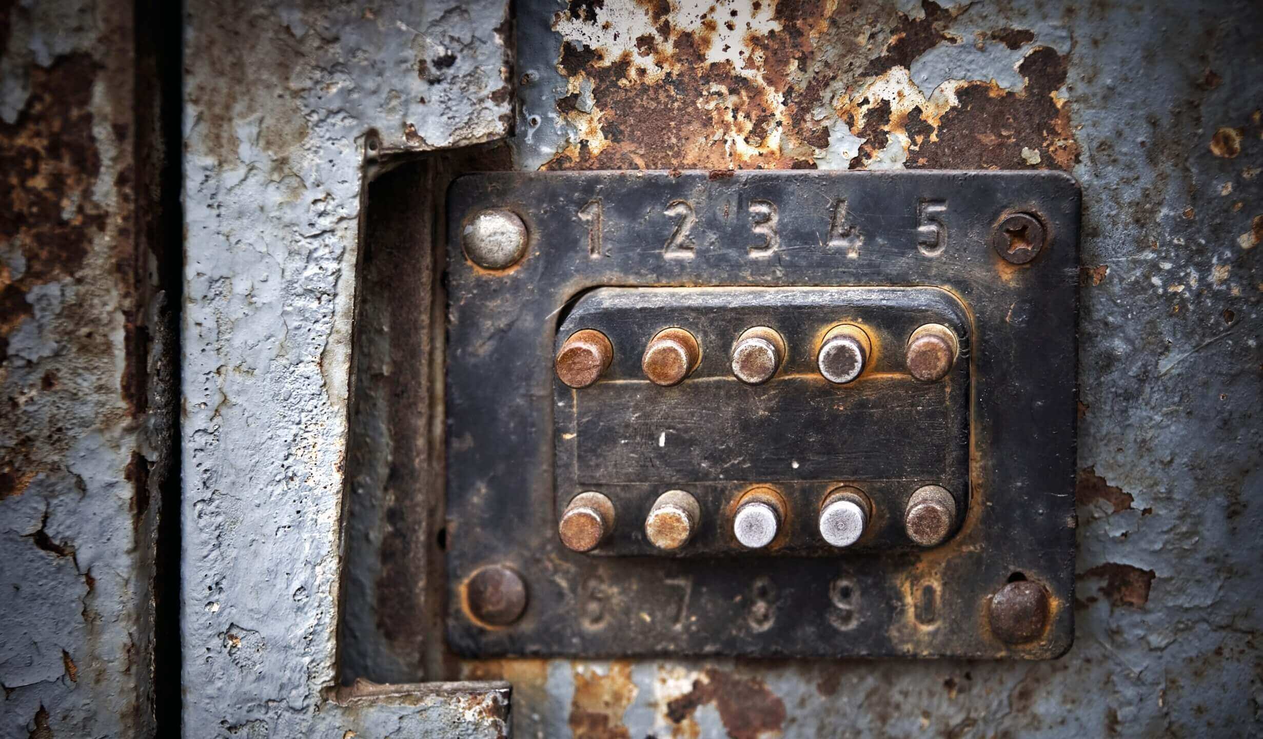 Detailaufnahme eines alten, rostigen Zahlenschlosses mit Metallknöpfen und sichtbaren Nummern auf verwittertem Untergrund